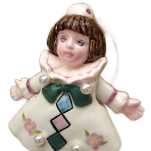 May include: A porcelain figurine of a clown with a white dress, green and pink diamond pattern, and a green bow. The clown has brown hair, blue eyes, and a red dot on its hat.