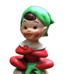 May include: A ceramic elf figurine with a green hat, red and green clothing, and a white base with yellow dots. The elf is sitting with its arms crossed and has a smiling face.