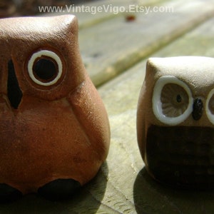 May include: Two ceramic owl figurines. The larger owl is brown with black eyes and a white eye outline. The smaller owl is tan with black and white details. The owls are on a wooden surface.