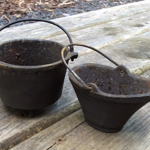 May include: Two vintage cast iron buckets with wire handles. The buckets are black and have a rustic, aged appearance.