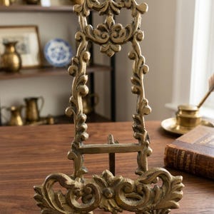 May include: Ornate gold-colored metal easel with a decorative, scrolling design. The easel has a three-legged base and a top with a circular flourish. It is designed to hold a small picture or plate. The style is vintage and antique.