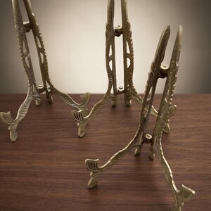 May include: Three brass display stands featuring an ornate, textured metal design with a hinged construction. Each stand has two curved legs at the base and a tall, slender frame with decorative detailing, designed to hold items upright.