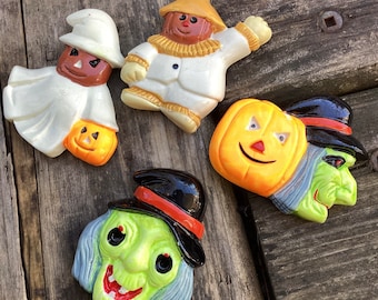 May include: Four Halloween-themed ceramic figurines. A ghost figure holding a jack-o'-lantern, a scarecrow, a smiling jack-o'-lantern, and a green-faced witch wearing a black hat with an orange band. The figurines are painted in bright colors.