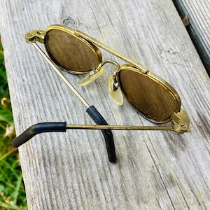 May include: Vintage gold-tone sunglasses with brown lenses. The frames have a double bridge design and black temple tips. The lenses are oval-shaped. The sunglasses are resting on a weathered wooden surface.