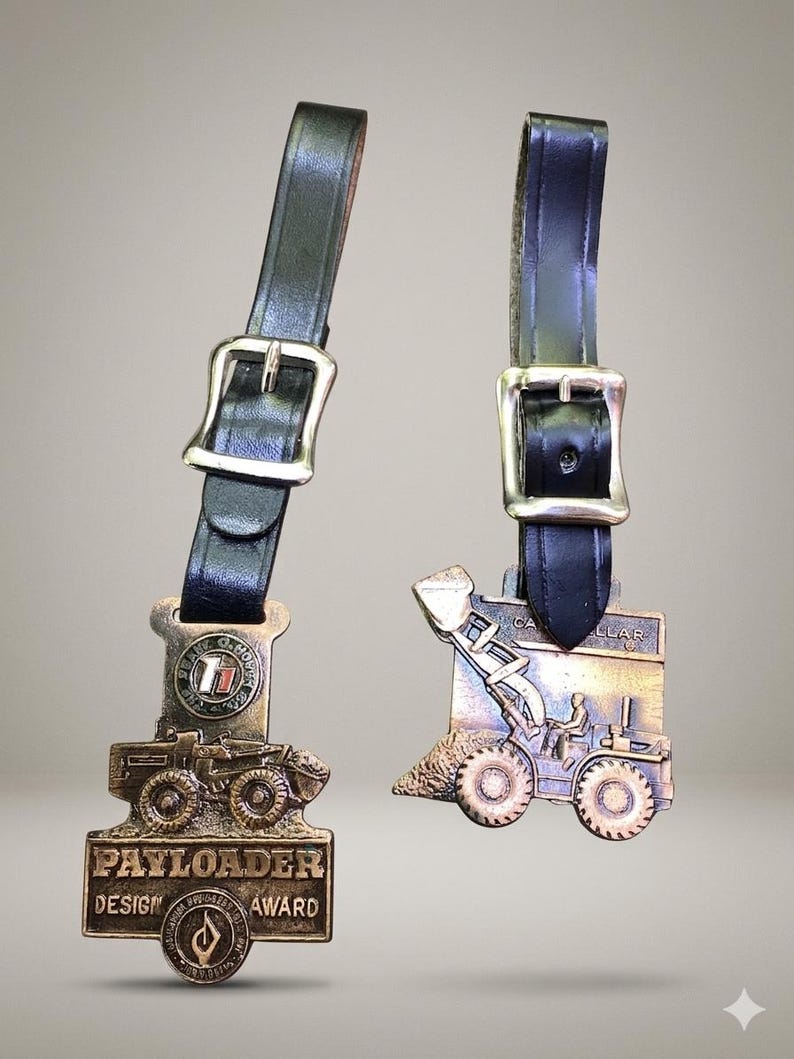 May include: Two bronze-toned metal award plaques attached to black leather straps with silver-colored metal buckles. The left plaque features a construction vehicle and the text PAYLOADER DESIGN AWARD. The right plaque depicts a Caterpillar loader.