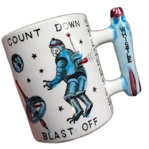 Peut inclure: Une tasse en céramique blanche avec une illustration d'un astronaute bleu et le texte "COUNT DOWN BLAST OFF". La poignée est en forme de fusée et porte l'inscription "WHISTLE".