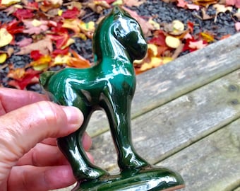 May include: A green ceramic horse figurine with a glossy finish. The figurine is standing on a wooden surface.