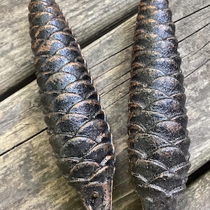 May include: Two elongated, cone-shaped decorative objects made of dark, textured metal with a bronze-toned finish on the edges of the scales. Each piece features a small, circular hole at the bottom end, designed for hanging or attachment purposes.