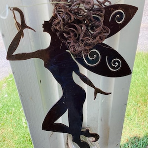 May include: A dark metal fairy silhouette with curly hair and decorative wings. The fairy is holding a small object and is positioned on a wooden surface. The background is a white structure with green grass visible.