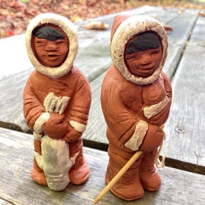 May include: Two terracotta-colored Inuit figurines with white-trimmed hoods and parkas. One holds a white object, the other a wooden stick. The figures have black hair and are standing on a wooden surface. The figurines are handmade.