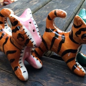 May include: Two ceramic tiger salt and pepper shakers. The salt shaker is pink, white, and orange with black stripes. The pepper shaker is green, white, and orange with black stripes.