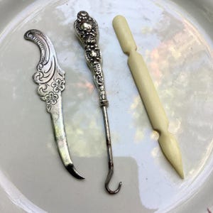 May include: A set of three vintage tools for buttoning and sewing. The tools are made of silver and bone. The silver tool has a decorative handle and a hook on the end. The bone tool is a long, thin needle with a pointed tip.