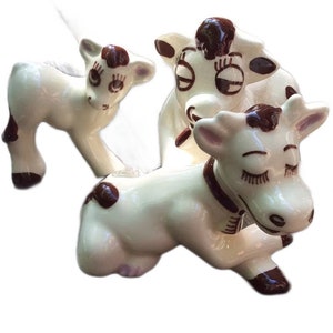 May include: Three ceramic cow figurines with brown spots and closed eyes. The cows are white with brown accents and are in a playful pose.