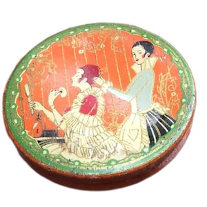 Puede incluir: Una lata redonda de color naranja con un borde verde y dorado. La lata presenta una ilustración vintage de dos mujeres, una aplicándose maquillaje mientras la otra observa.