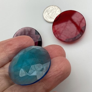 May include: A selection of colourful, faceted glass cabochons in teal, red, and purple. The cabochons are round and oval, with a coin for scale. The teal cabochon is held in a hand.