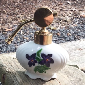 Peut inclure: Un flacon de parfum en porcelaine blanche avec un atomiseur en métal doré et un motif floral représentant des violettes violettes. Le flacon a un bord festonné et un design ondulé.