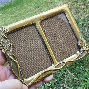 May include: A vintage brass double photo frame with floral detailing. The frame is rectangular with two openings and a textured backing. The floral design is on the corners and sides of the frame.