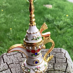 May include: A decorative white and gold teapot with jewel accents. The teapot features a long, ornate spout and handle, with a pearl-like top. The design includes red, blue, and green jewels, and floral patterns. The teapot is set against a blurred green background.