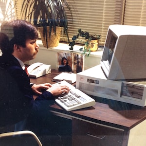 May include: A person is seated at a desk with a computer, keyboard, and telephone. They are wearing a dark suit and tie. The desk is made of wood and has a drawer. There is a plant in a pot on the desk.