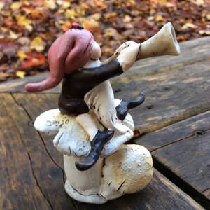 Op de afbeelding: Een grillig keramiek beeldje van een kabouter die op een hoorn blaast, zittend op paddenstoelen. De kabouter draagt een bruine jas, witte broek en zwarte laarzen. Op zijn hoofd zit een rode kever. De paddenstoelen zijn wit met een textuur.