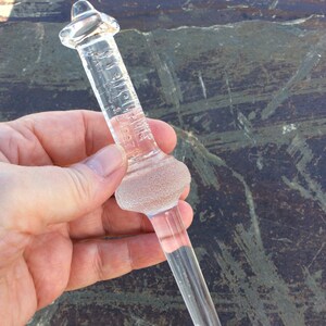 May include: A clear glass dropper bottle with a textured bulb and a long, thin neck. The bottle is marked with the words "1/2 FLUID DRAM" and "1/8 OZ."