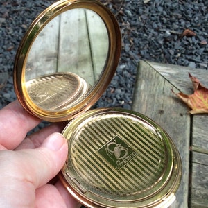 May include: A gold-toned compact mirror with a striped design and the inscription "Stratton" on the lid. The mirror is open and the reflection of a wooden surface is visible.