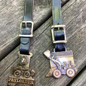 May include: Two bronze-toned metal award plaques attached to black leather straps with silver-colored metal buckles. The left plaque features a Payloader tractor and text reading PAYLOADER DESIGN AWARD. The right plaque depicts a Caterpillar tractor.