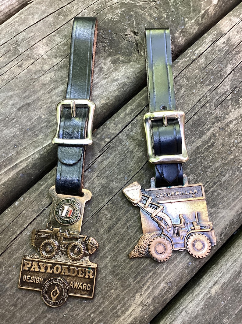 May include: Two bronze-toned metal award plaques attached to black leather straps with silver-colored metal buckles. The left plaque features a Payloader tractor and text reading PAYLOADER DESIGN AWARD. The right plaque depicts a Caterpillar tractor.
