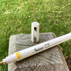 May include: A white pen with a black label that says "Stano CS" and a yellow number "0" on the side. The pen is next to a white cap with the word "Standard" printed on it.