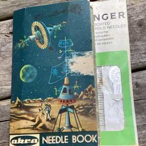 May include: Vintage "Akra Needle Book" with a space-themed illustration. The cover depicts a rocket, astronaut, and planets against a dark blue sky. Next to it is a green and white package of "Singer" sorted hold needles.