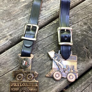 May include: Two bronze-toned metal award plaques featuring construction vehicle designs, each attached to a black leather strap with a silver-colored metal buckle. The left plaque reads PAYLOADER DESIGN AWARD and THE FRANK G. HOUGH CO, while the right reads CATERPILLAR.