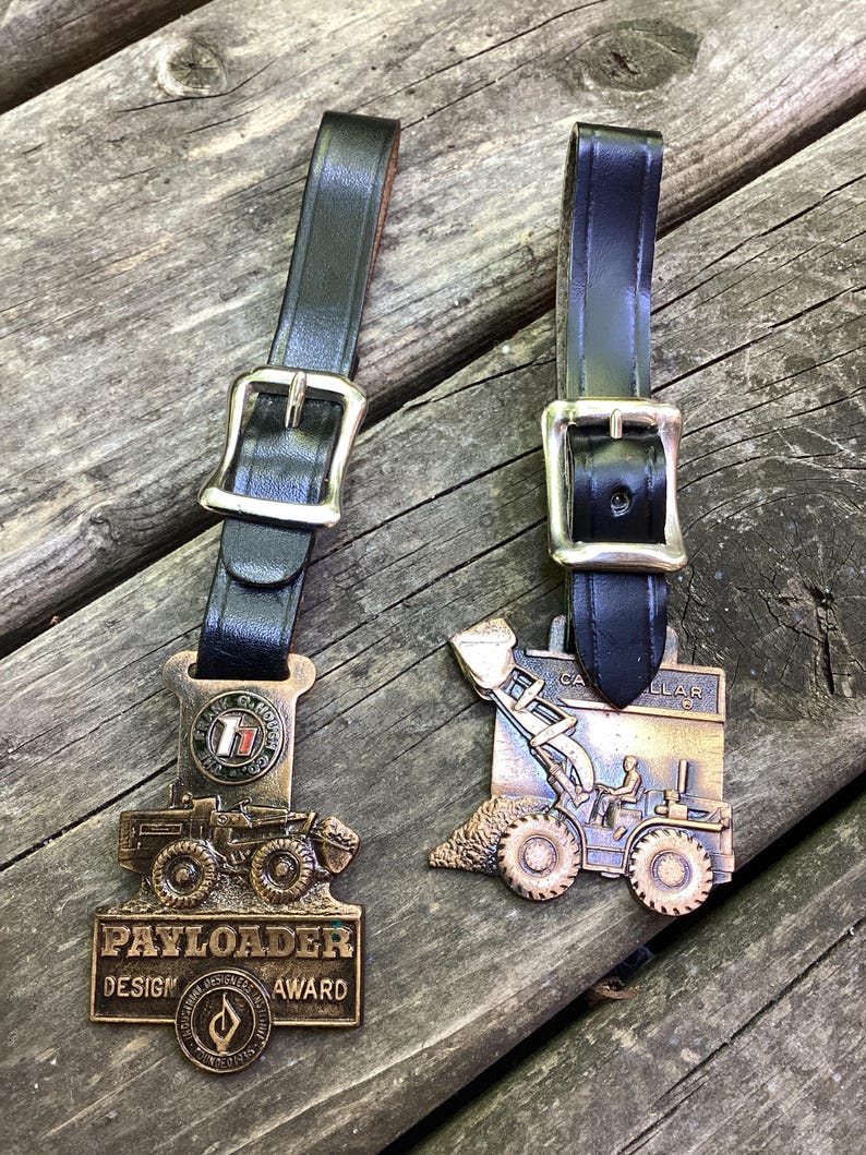 May include: Two bronze-toned metal award plaques featuring construction vehicle designs, each attached to a black leather strap with a silver-colored metal buckle. The left plaque reads PAYLOADER DESIGN AWARD and THE FRANK G. HOUGH CO, while the right reads CATERPILLAR.