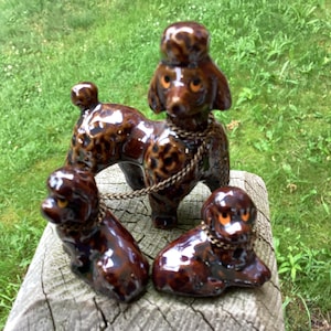 May include: Three brown ceramic poodle figurines. One standing poodle is connected to two smaller, seated poodles by a gold-colored chain. The poodles have orange eyes and a glossy finish. The figurines are on a wooden surface.