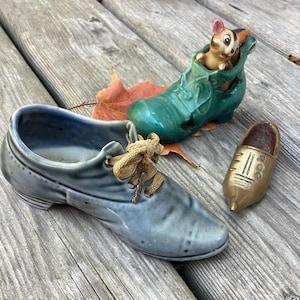 May include: Three ceramic shoe figurines. A blue shoe with tan laces, a green shoe with a mouse figurine inside, and a small gold shoe. The figurines are on a wooden surface.