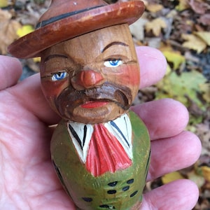 May include: A wooden figurine of a man wearing a brown hat and a green jacket with red and white accents. The figurine has a painted face with blue eyes and a moustache.