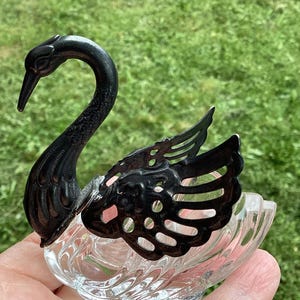 May include: A decorative swan-shaped dish with a black metal swan head and wings. The dish is made of clear glass with a textured, scalloped design. The swan's head is gracefully curved, and the wings have an open, ornate pattern.