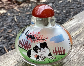 May include: A clear glass bottle with a red lid and a painted design of a black and white dog in a garden setting. The bottle has a white background with red, blue, and green accents.