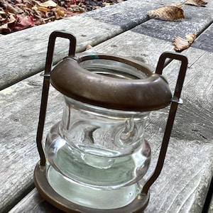 Peut inclure: Une lanterne en verre transparent avec un cadre et une poignée en métal couleur laiton. La lanterne a une base ronde et un dessus large. Le verre est texturé et la lanterne est posée sur une surface en bois.