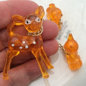 May include: Orange plastic deer charm with white polka dots and a gold chain. The deer is attached to a smaller orange plastic charm with a gold chain.