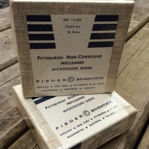 May include: Two vintage boxes of Pittsburgh Non-Corrosive Precleaned Microscope Slides. Each box is beige with black text and graphics, including the Fisher Scientific logo. The boxes are labeled with dimensions of 75x25 mm.