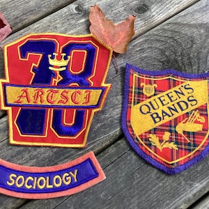 May include: Three embroidered fabric patches. One large red patch features the blue text 79, a yellow crown, and the yellow text ARTSCI. A shield-shaped patch has a red plaid pattern, yellow banner with blue text QUEEN'S BANDS, and a small yellow harp. A curved patch has a blue centre with yellow text SOCIOLOGY.