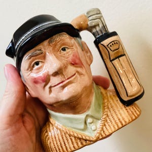 May include: Ceramic golf-themed mug shaped like a smiling man wearing a black hat and a tan sweater. The handle is a golf club bag with golf clubs. The man has rosy cheeks and a green collared shirt.