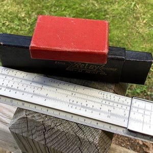 May include: A vintage slide rule with a metal body, resting on a wooden surface. A black case with the text "Relay Keiki Co." and a red box are also present. The slide rule is a tool for mathematical calculations.