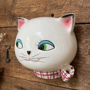 May include: A white ceramic cat head wall hanging with pink-lined ears, blue eyes, and a red and white checkered scarf. The cat has black whiskers and a black nose. The cat is mounted on a wooden surface.
