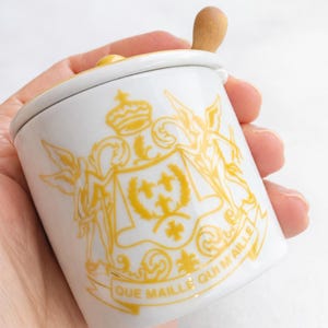May include: White ceramic jar with a lid and a small wooden spoon. The jar features a yellow crest design with the words "QUE MAILLE QUI M'AILLE" in yellow. The jar is held in a hand.