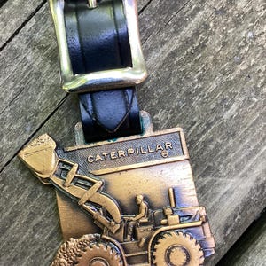May include: A bronze-toned metal tag featuring a relief of a Caterpillar construction vehicle with a raised bucket and operator, attached to a black leather strap with a silver-colored metal buckle. The word CATERPILLAR is embossed at the top.