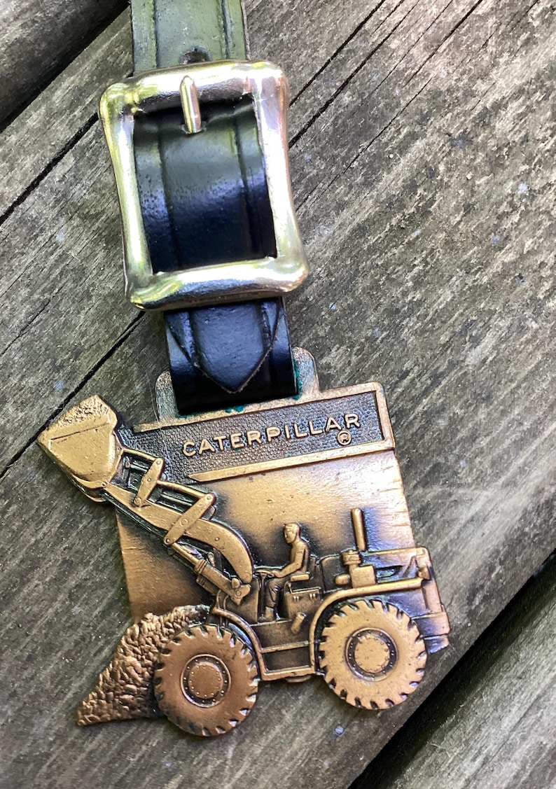 May include: A bronze-toned metal tag featuring a relief of a Caterpillar construction vehicle with a raised bucket and operator, attached to a black leather strap with a silver-colored metal buckle. The word CATERPILLAR is embossed at the top.