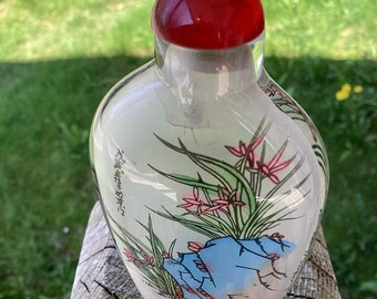 May include: A clear glass perfume bottle with a red stopper. The bottle is decorated with a painted floral design in pink, green, and blue. The bottle has a rounded shape and is sitting on a wooden surface.