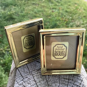 May include: Two square, gold-coloured brass picture frames. Each frame has a label that reads "hand polished SOLID BRASS". The frames are on a weathered wooden surface, with a blurred green background.