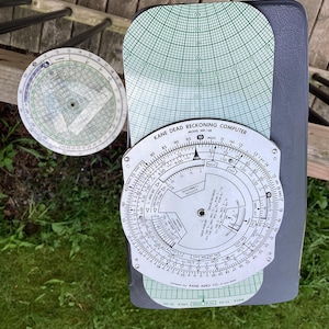 May include: A silver circular dead reckoning computer with a green and white gridded chart. The computer has a dial with markings for miles, knots, and altitude. The text "KANE DEAD RECKONING COMPUTER MODEL MK-68" is printed on the chart.